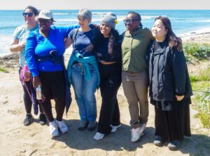 Six women stand on a dune with the Pacific Ocean behind them.